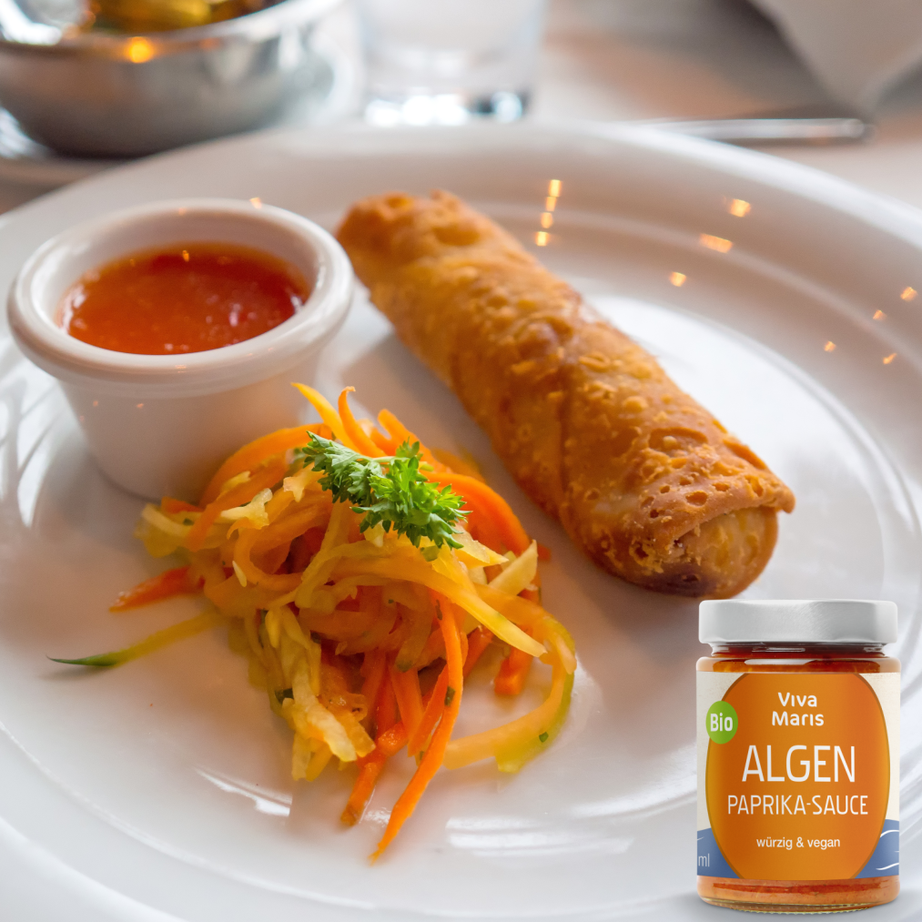 Viva Maris Bio Algen Paprika Sauce im Glas vor Teller mit Salat und Frühlingsrolle und einem Pot Sauce